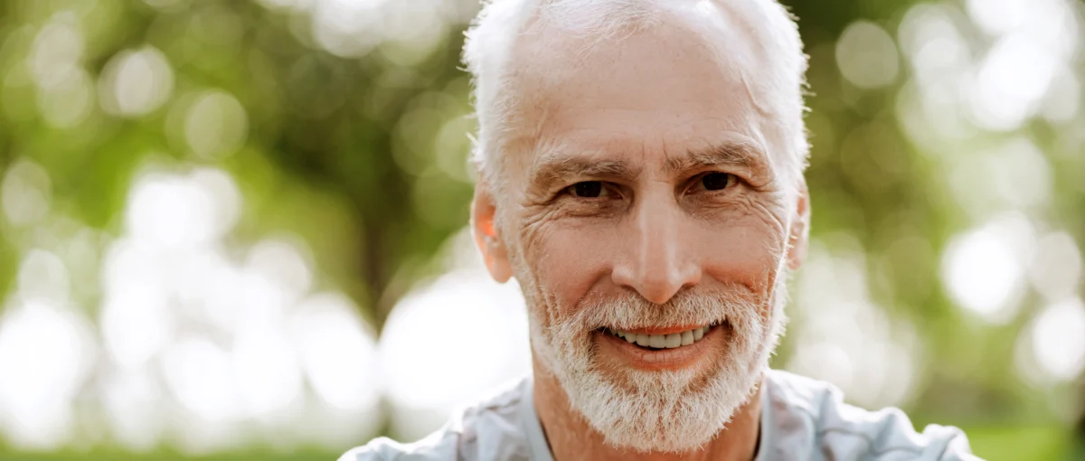 A man sitting and smiling while surrounded by trees in sunlight.