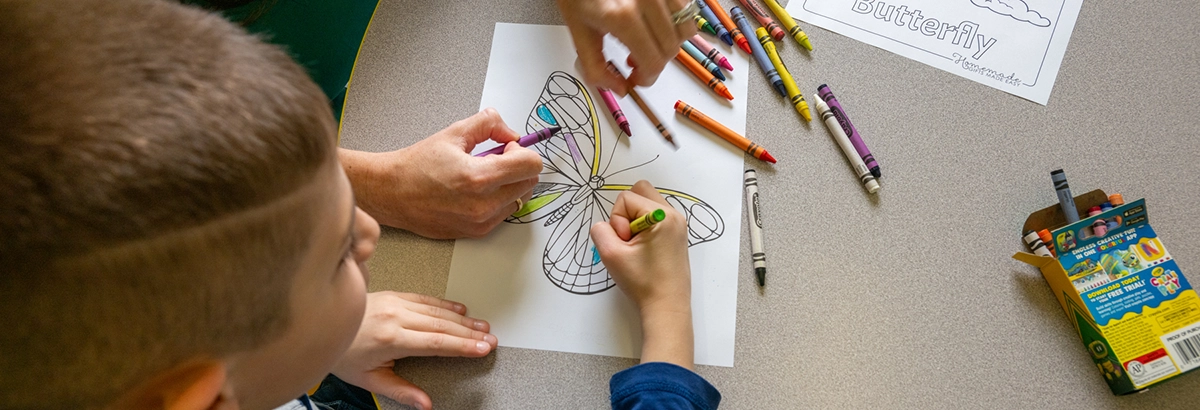 Aerial view of a child coloring in a butterfly at a table with crayons scattered around.