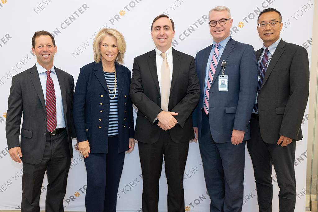 A group of smiling professionals posing in front of a New York Proton Center backdrop at the center's 5th Anniversary event.