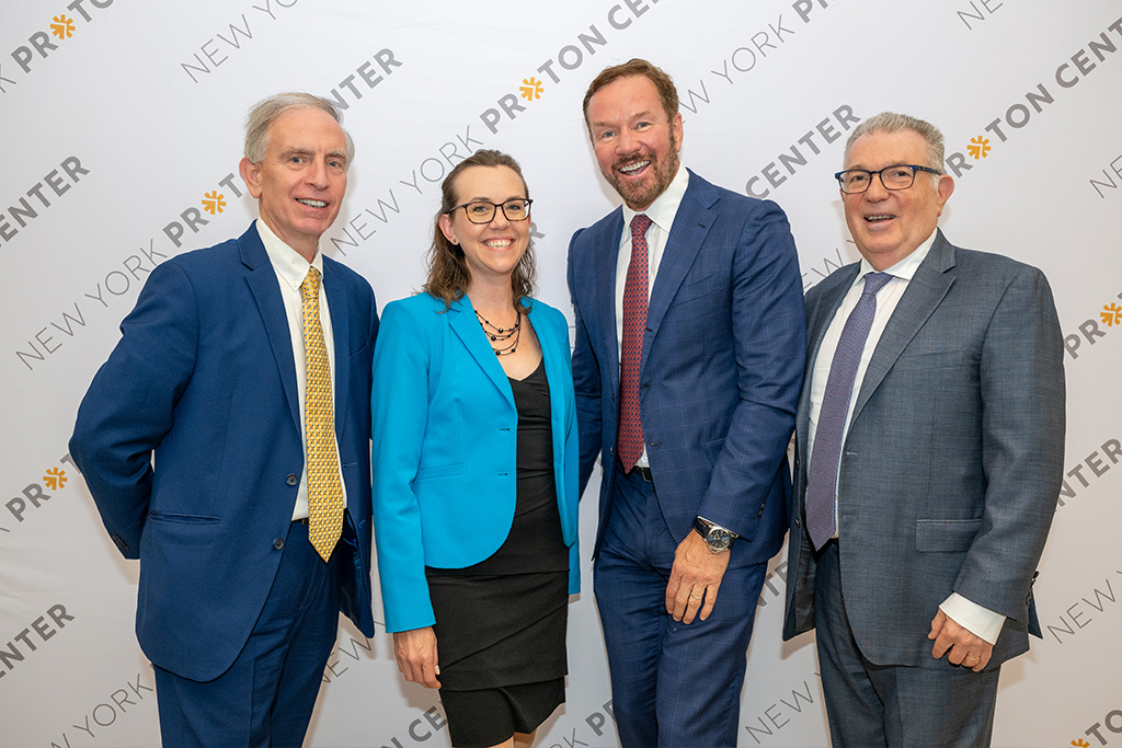 A group of four smiling professionals posing in front of a New York Proton Center backdrop at the center's 5th Anniversary event.