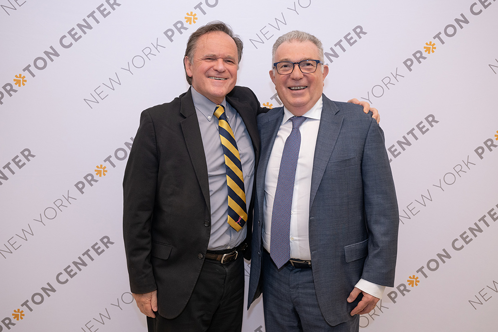 Two men in suit and ties standing and smiling in front of a New York Proton Center branded backdrop.