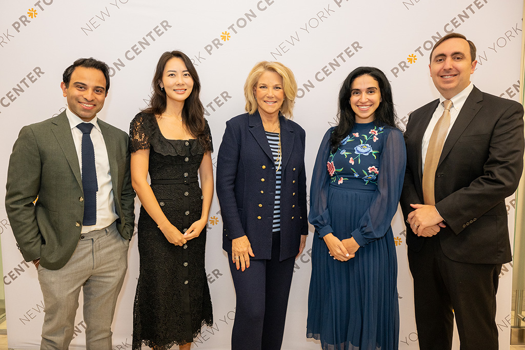 Three women standing between two men in formal attire posing as a group and smiling in front of a New York Proton Center branded backdrop.