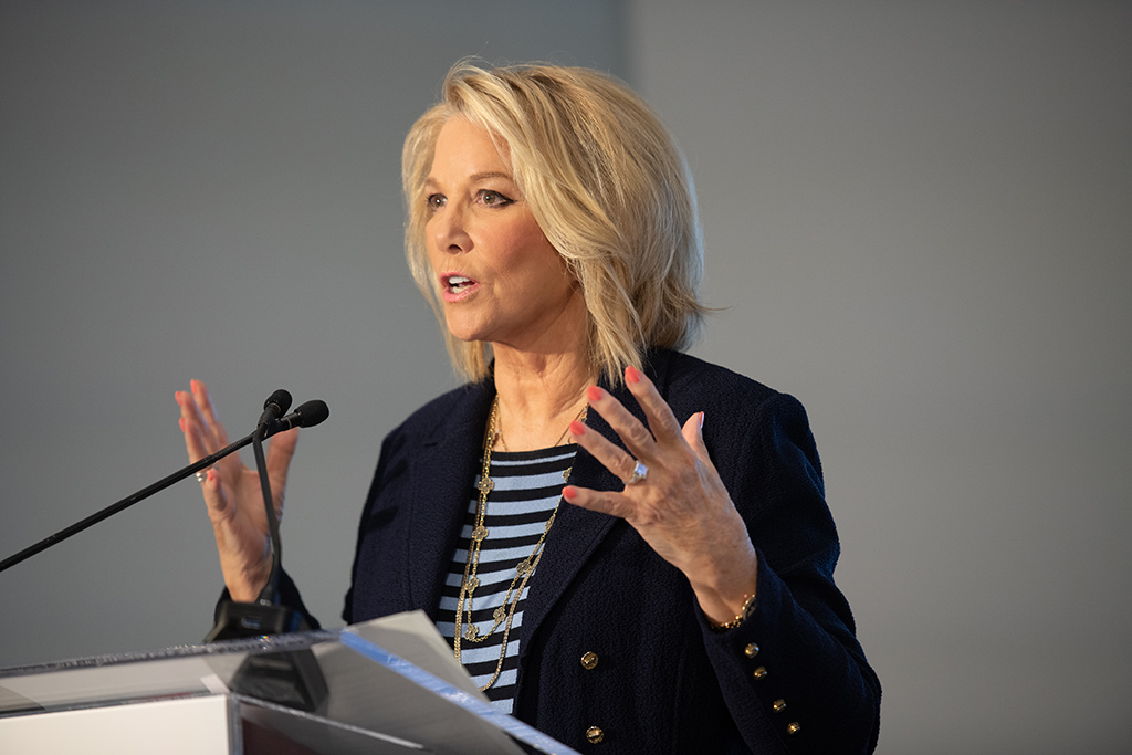Woman with blonde hair speaks passionately into a microphone on a podium.
