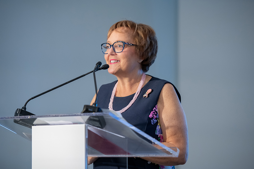 Woman with glasses on speaks into a microphone on a podium.