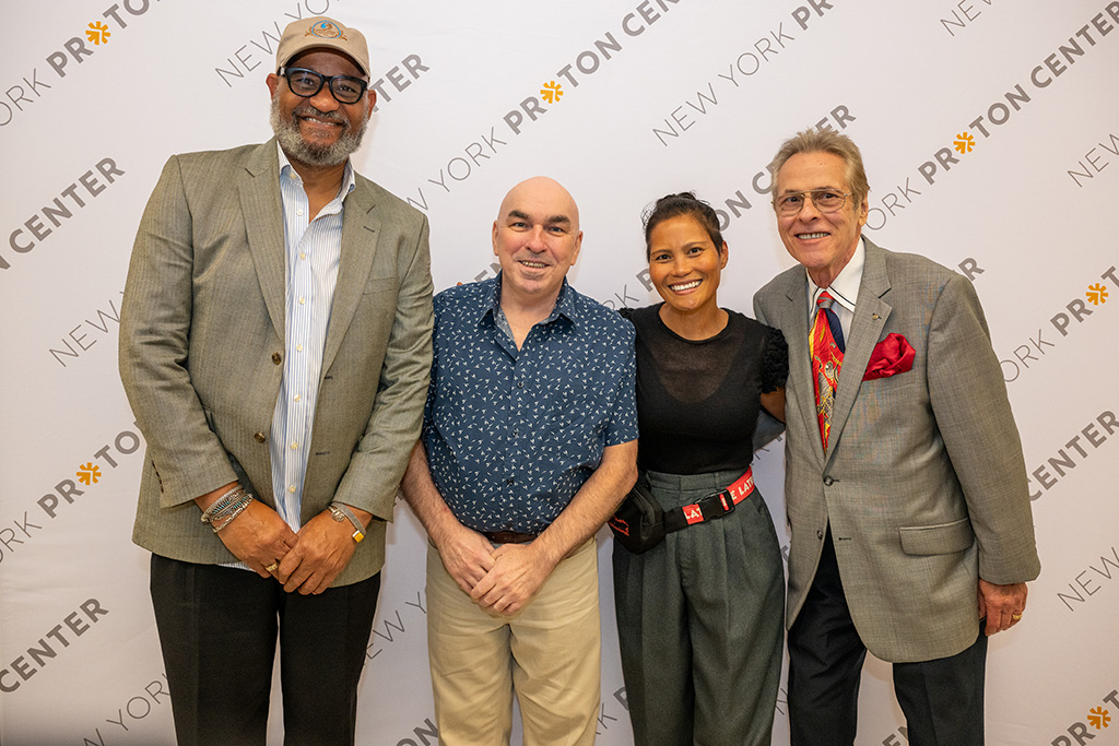 Three men and one woman standing as a group smiling in front of a New York Proton Center branded backdrop.