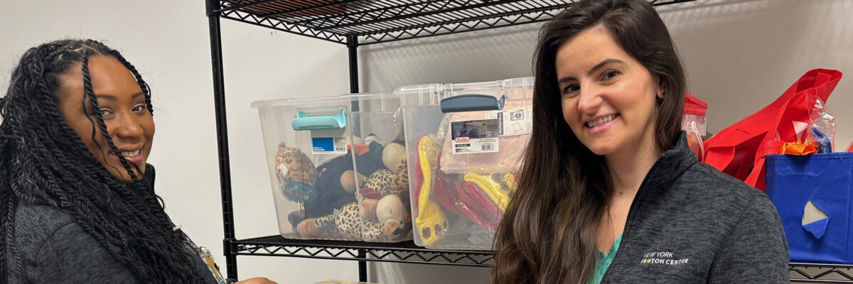 Two New York Proton Center staff smiling among bins of donated toys during their annual toy drive.
