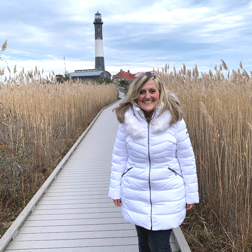 NYPC patient, Debi Walsh Lembo, standing on a scenic wooden boardwalk with a lighthouse in the background happily smiling.