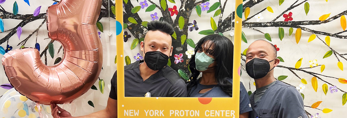 Three masked New York Proton Center nurses huddled together posing with a selfie frame and a number 3 balloon inside the center