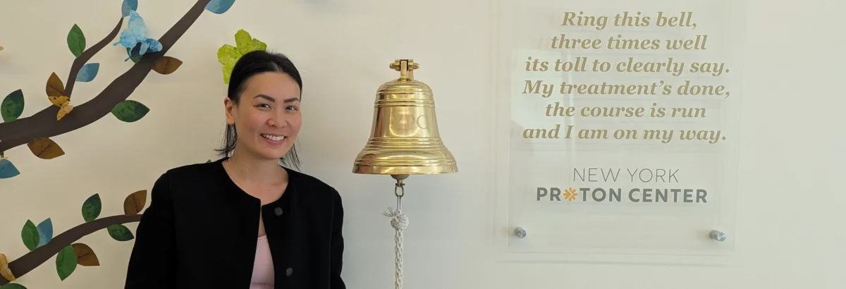 New York Proton Center patient Irene smiles while standing next to an end of cancer treatment bell.