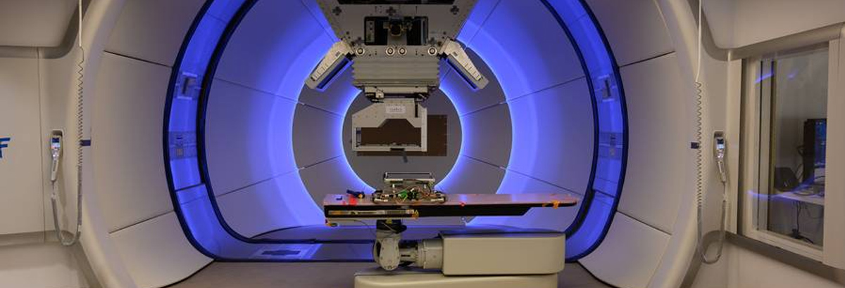 A proton therapy treatment room with treatment table surrounded by rotating equipment against a blue-lit background.