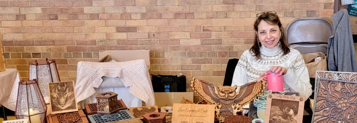 Woman with brown hair smiling and sitting at a table of wooden arts and crafts.